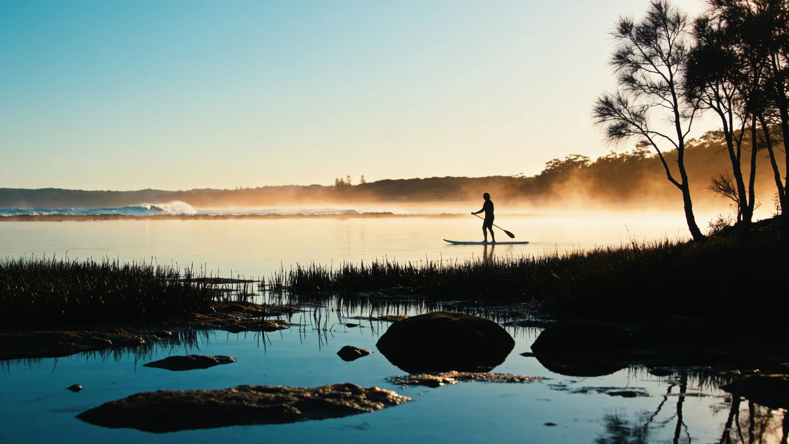 Early morning light over Narrabeen Lagoon, with mist lifting off the water and a lone paddleboarder crossing the still surface.